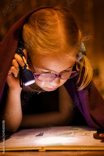 Little blonde girl 7 years old with glasses reading the book under the blanket with little flashlight at dark night time photo