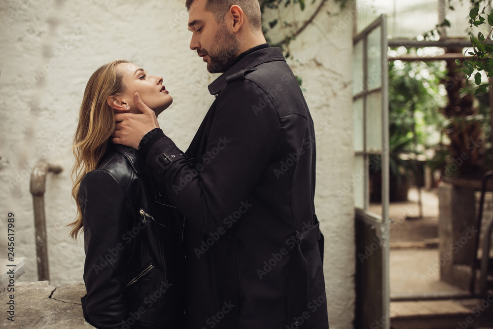 Young handsome man in total black clothes holding hand on neck of pretty woman dreamily looking at each other while spending time together in old cozy greenhouse Young handsome man in total black clothes holding hand on neck of pretty woman dreamily looking at each other while spending time together in old cozy greenhouse