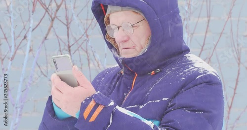 Elderly man with glasses holds smartphone in his hands. Pensioner in blue warm jacket in courtyard rural house in background of landscape with ship. Bad weather, storm warning, village, winter, snow