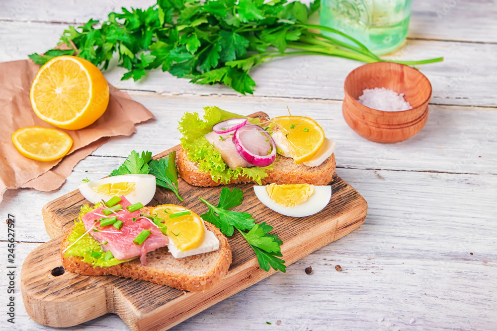 Sandwich with herring and tuna fillets with fresh green salad, onion ring, a quarter of eggs, home cheese, piece of lemon, micro greens and scallions on the wooden desk Sandwich with herring and tuna fillets with fresh green salad, onion ring, a quarter of eggs, home cheese, piece of lemon, micro greens and scallions on the wooden desk