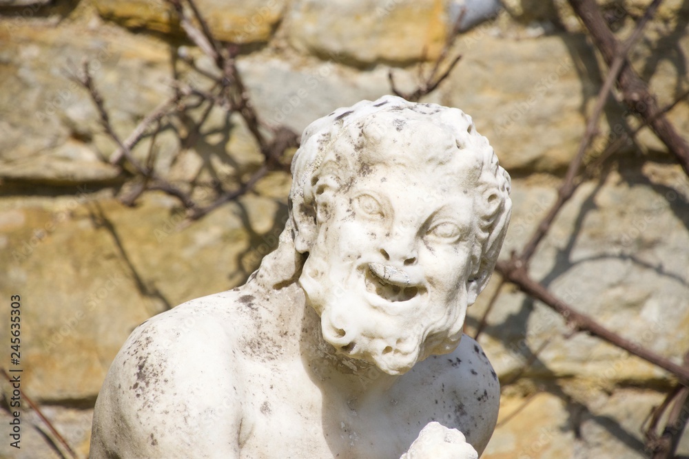 A stone statute, outdoors and in bright sunlight, of a funny old man with a shrivelled face and a big laughing grin that makes him look like a satyr or goblin. Curly hair and beard; possibly evil.