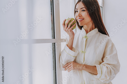 Happy young girl tasting apple near wide window