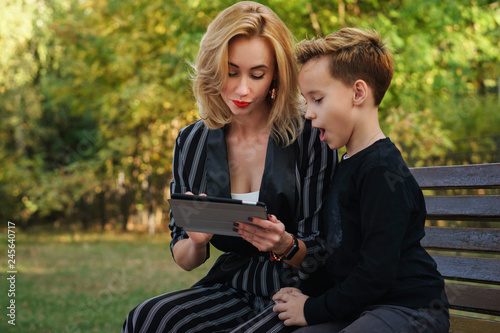 Young attractive mother and son are sitting on bench in city park. Woman is showing something on tablet pc screen. Boy is very surprised. Family time © Vagengeim