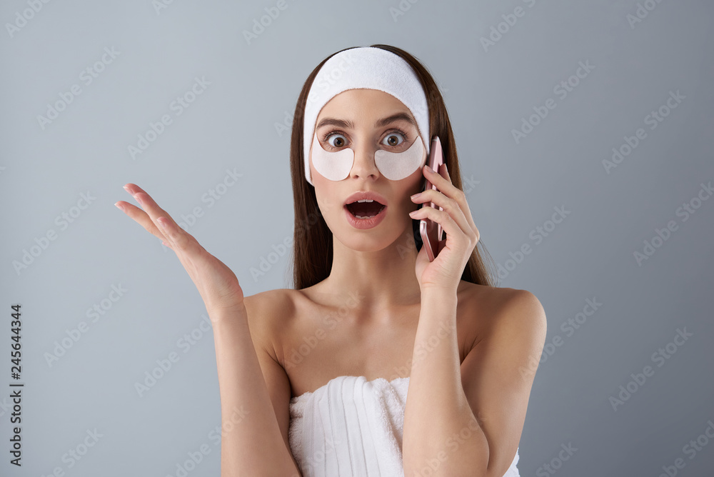 Astonished young lady with under-eye patches talking on cellphone Astonished young lady with under-eye patches talking on cellphone