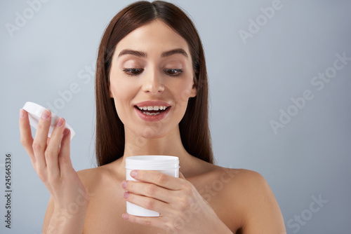 Cheerful young lady opening white jar of cosmetic cream
