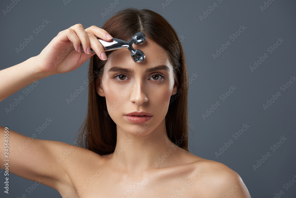 Beautiful young lady with naked shoulders using facial massager Beautiful young lady with naked shoulders using facial massager