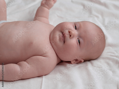 Baby lying down playfully looks into the camera. The baby lies on a white linen cloth.