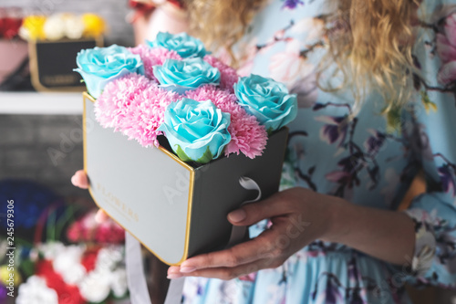 Flower Workshop: Florist girl is holding a small box with a bouquet of carnations and roses. Around on the shelves are many colorful bright bouquets.