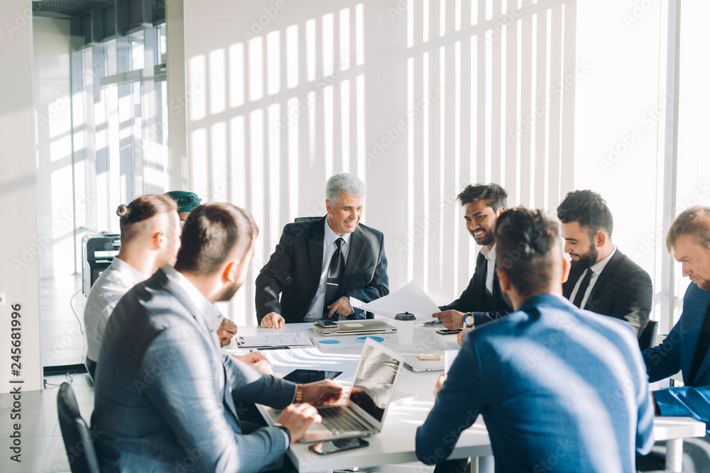 Multiracial male business executives discussing project sitting at conference table in spacious hall with panoramic city scapes, all men dressed in formalwear and have positive mood Multiracial male business executives discussing project sitting at conference table in spacious hall with panoramic city scapes, all men dressed in formalwear and have positive mood
