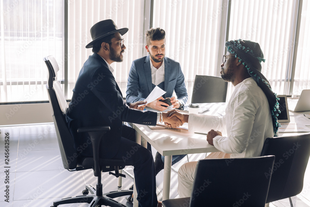Two mixed race executives men are sitting at the desk and shaking hands while caucasian male assistant is looking at them and smiling Two mixed race executives men are sitting at the desk and shaking hands while caucasian male assistant is looking at them and smiling