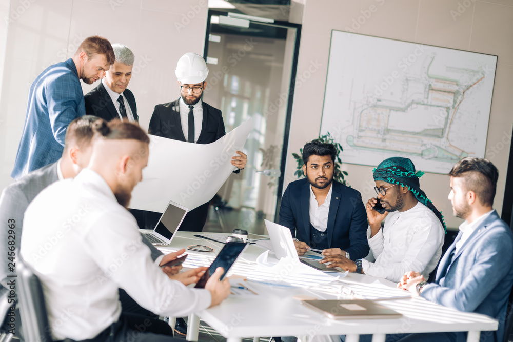 Multiracial group of constructors, builders, engeneers and architects discussing blueprint at office. All men are dressed in business suits, and white shirts, one of them wears hardhat on head