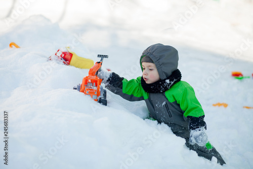 Portrait of cute little toddler sitting on snow and playing with his yellow tractor toy in the park. Child playing outdoors. Happy boy with construction toy. Lifestyle concept
