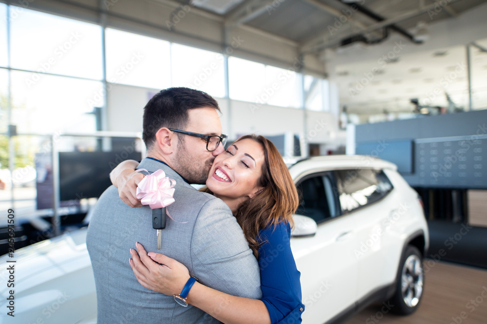 Lovely couple celebrating their car purchase at vehicle dealership showroom. Woman hugging his man holding car keys in front SUV vehicle. Lovely couple celebrating their car purchase at vehicle dealership showroom. Woman hugging his man holding car keys in front SUV vehicle.