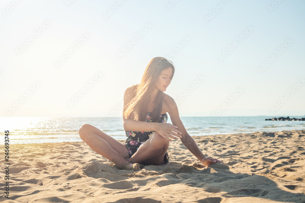 A girl sits on a sandy beach and runs her hand through the sand.