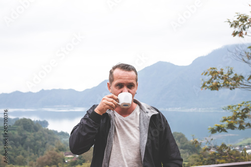 Young man traveler with white cup of coffee on a mountain background. Bali island.