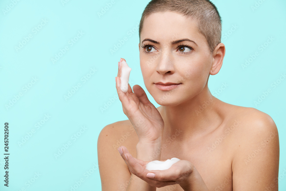 Smiling mid 30s woman using gentle foam facial cleanser. Photo of attractive caucasian woman with shaved head and healthy skin isolated on pastel blue background washing her face with foam soap. Smiling mid 30s woman using gentle foam facial cleanser. Photo of attractive caucasian woman with shaved head and healthy skin isolated on pastel blue background washing her face with foam soap.