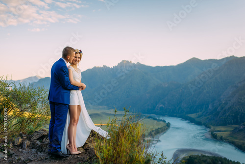 Young newly wed couple, bride and groom kissing, hugging on perfect view of mountains, river and blue sky