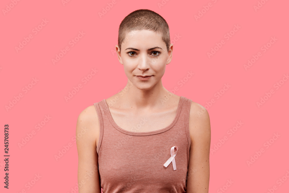 Smiling mid 30s woman, a cancer survivor, wearing pink breast cancer awareness ribbon, isolated over living coral background. Support, solidarity, screening and prevention concept. Smiling mid 30s woman, a cancer survivor, wearing pink breast cancer awareness ribbon, isolated over living coral background. Support, solidarity, screening and prevention concept.