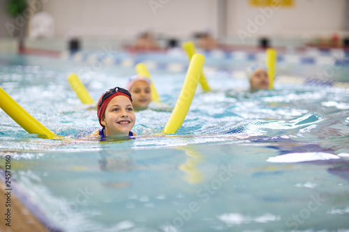 Happy little girl in swimming cap learning to swim with swim noodle at the lesson in pool