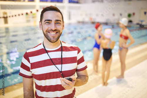 Portrait of happy young swim instructor holding stopwatch and smiling with his class in the background