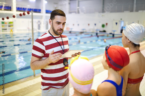 Young swim instructor standing with digital tablet and talking to the little girls before training in the swimming pool