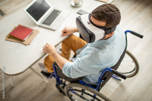 Disable man with vr headset sitting on wheelchair by desk with laptop and documents while working with virtual stuff
