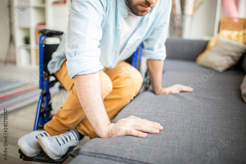 Young disable man trying to replace himself on sofa from wheelchair while sitting close to couch