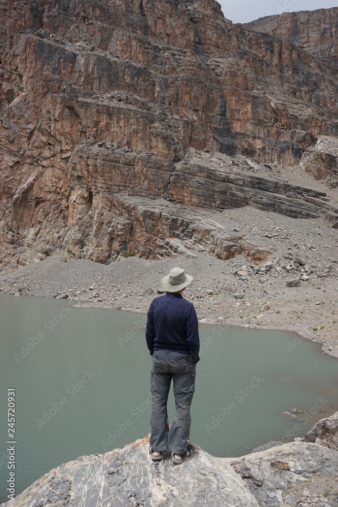 Man wearing a hat standing on the cliff looking at the mountain lake, Lake Mutnoe in Fann Mountains, Tajikistan 