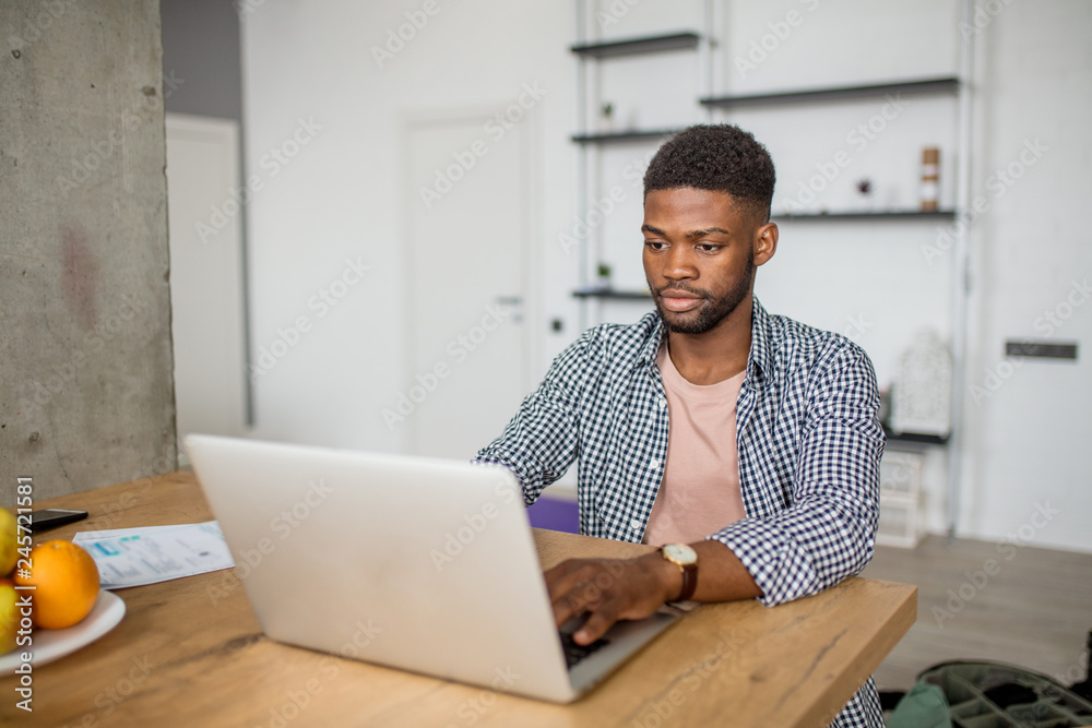 Young bearded African male blogger sitting at table in casual wear, writing article using his laptop at home Young bearded African male blogger sitting at table in casual wear, writing article using his laptop at home