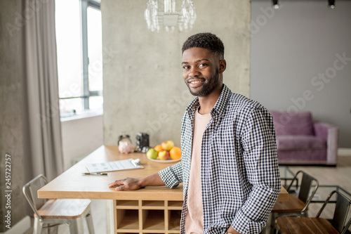 Happy young African bearded man in casual clothes looking at camera  posing over modern home interior.