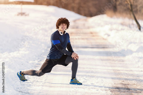 Smiling Caucasian man with curly hair and in sportswear stretching legs before running. Snow all around, healthy lifestyle concept.