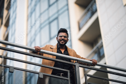 Young happy Indian businessman smiling with arms on handsrail at rooftop of business center photo