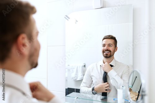 Smiling businessman putting on necktie while looking in the mirror and standing in the bathroom.