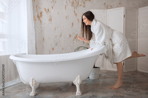 Beautiful young woman posing in bathrobe before taking bath, tempting with her slim fragile naked shoulders and long healthy hair.