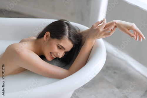 Beautiful bathing european brunette woman relaxing in milky bathtub  demonstrating her naked shoulder and arms