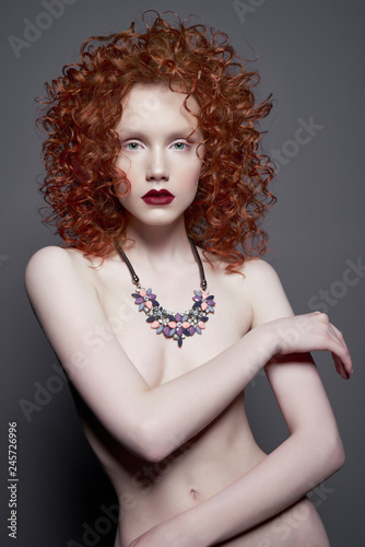 Beautiful young woman with black jewelry and red-hair on grey background. Nude pretty lady with bright red lipsstick and modern art makeup. Sensual model pose in photo-studio. Sexy naked body. photo