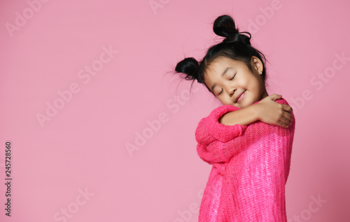 Asian kid girl with closed eyes in pink sweater hug herself and dream