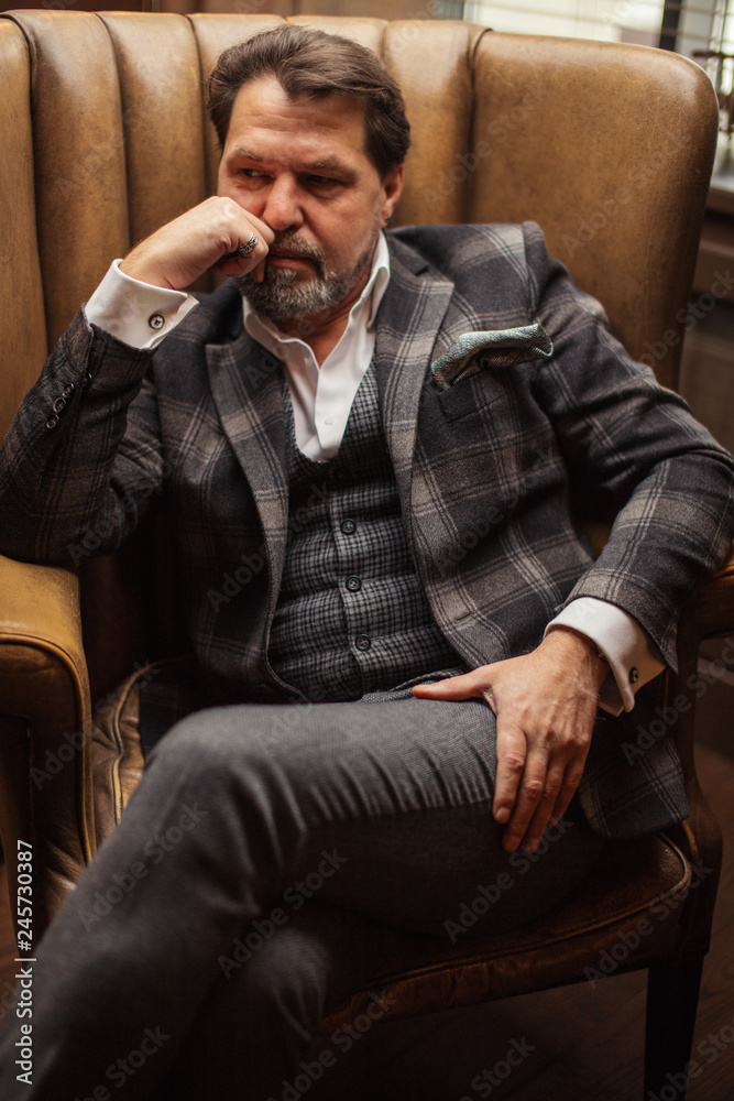 Pensive elegant bearded businessman thinks about something important while sitting in the leather armchair in living room Pensive elegant bearded businessman thinks about something important while sitting in the leather armchair in living room