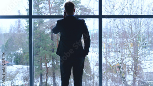A young guy (Man) businessman in suit is talking holding a telephone in the building (office) on the background of the street waiting for a meeting. Concept of: Nervous, Important Meeting.
