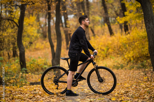 Handsome man biker in professional sportswear looking to side while riding bike down park alley on autumn day. Sportsman training thinking about future win in contest. 