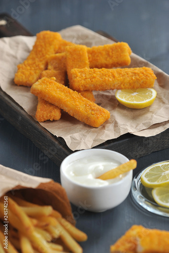 Fish sticks and french fries on a dark wooden background. French fries in a packet of Kraft paper. Sauce and lemon slices are used as a supplement. Close-up. Vertical frame orientation. 