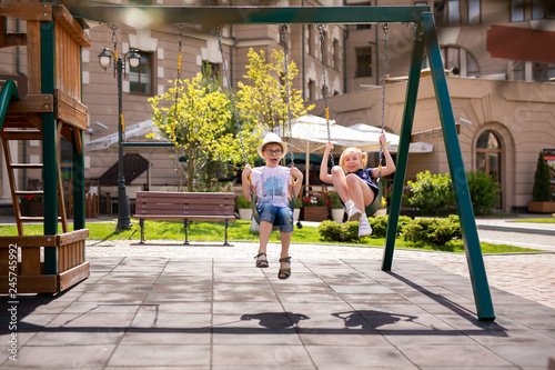 Boy in the glasses and hat and blonde girl in the dress having fun on a swing together in beautiful summer garden on warm and sunny day outdoors