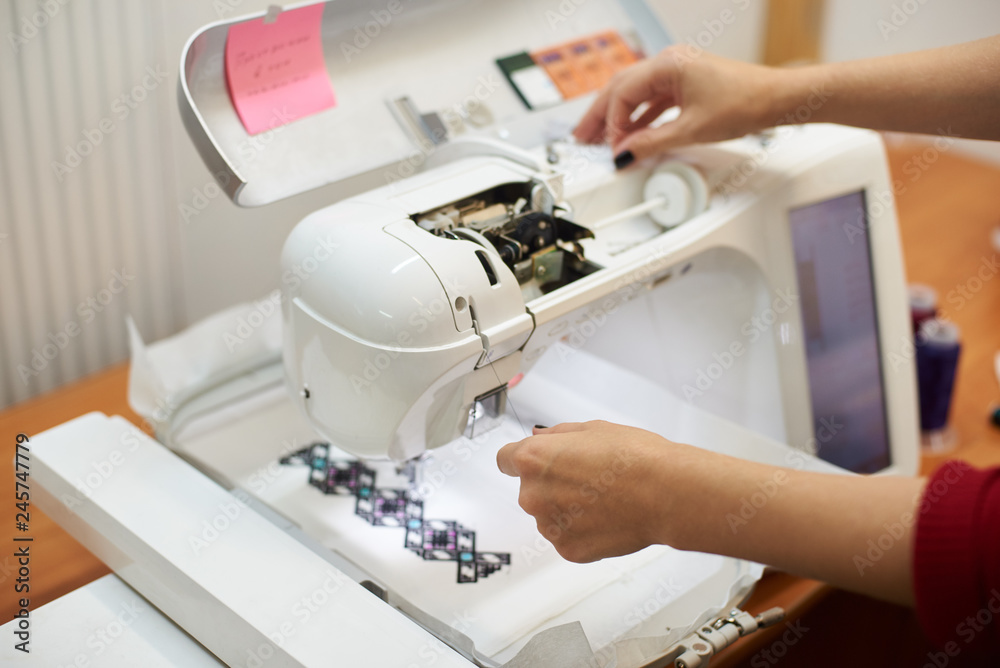Close-up of woman hands filling thread in sewing machine on background of white textile detail with embroidered colorful geometrical pattern. Modern technology and fashionable clothing manufacturing.