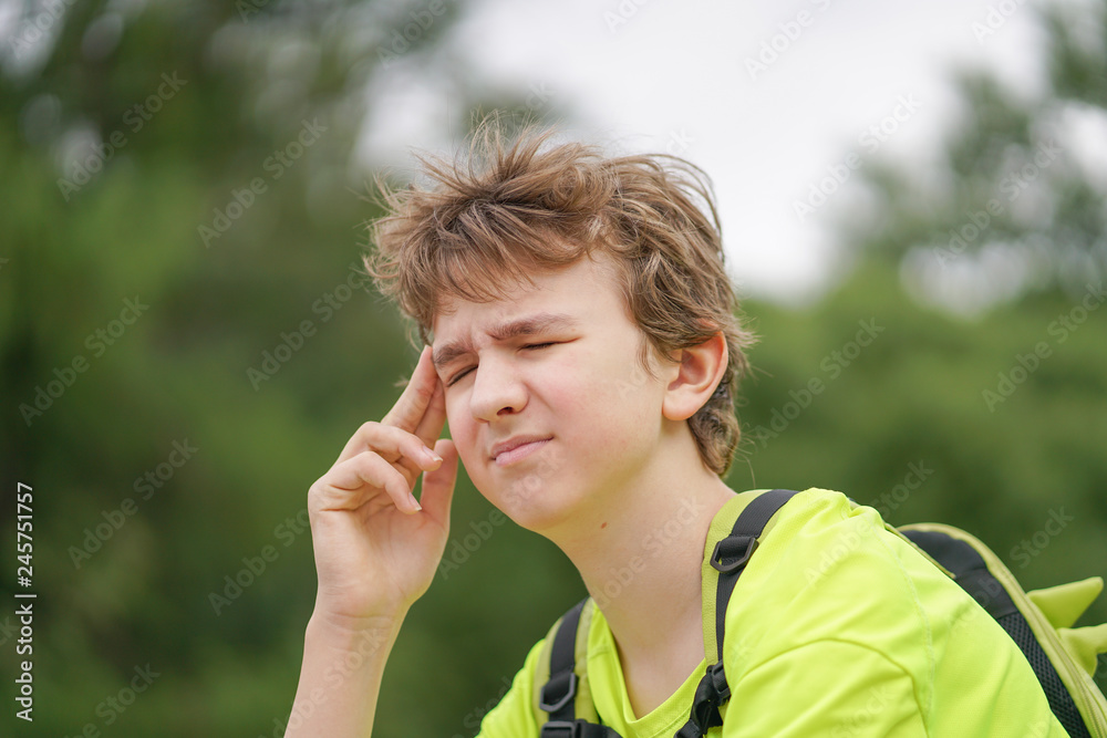 a young teenager guy is suffering from a headache. he keeps his hands to his head and winces of discomfort, sitting on the nature. a young teenager guy is suffering from a headache. he keeps his hands to his head and winces of discomfort, sitting on the nature.