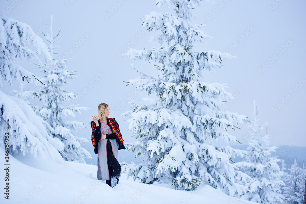 Attractive woman in short embroidered and decorated sleeveless fur coat made from sheepskin on snowy spruce trees blurred background. Traditional women's attire of western part of Ukraine.