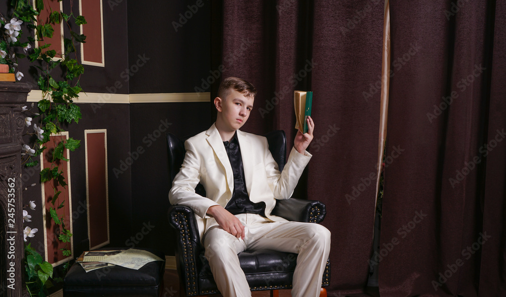 bored teen student guy in white business suit sitting in a chair with a book by the fireplace bored teen student guy in white business suit sitting in a chair with a book by the fireplace
