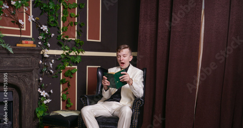 bored teen student guy in white business suit sitting in a chair with a book by the fireplace