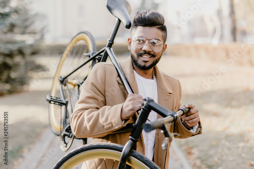 Bearded indian hipster man with a fixie bicycle. Confident young bearded man carrying his bicycle on shoulder and looking at camera while walking outdoors. Stylish businessman with a bike.