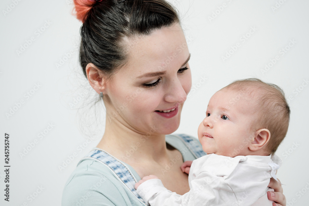 Young woman mother in denim overalls holds a baby child in her arms. White background in the studio. Young woman mother in denim overalls holds a baby child in her arms. White background in the studio.