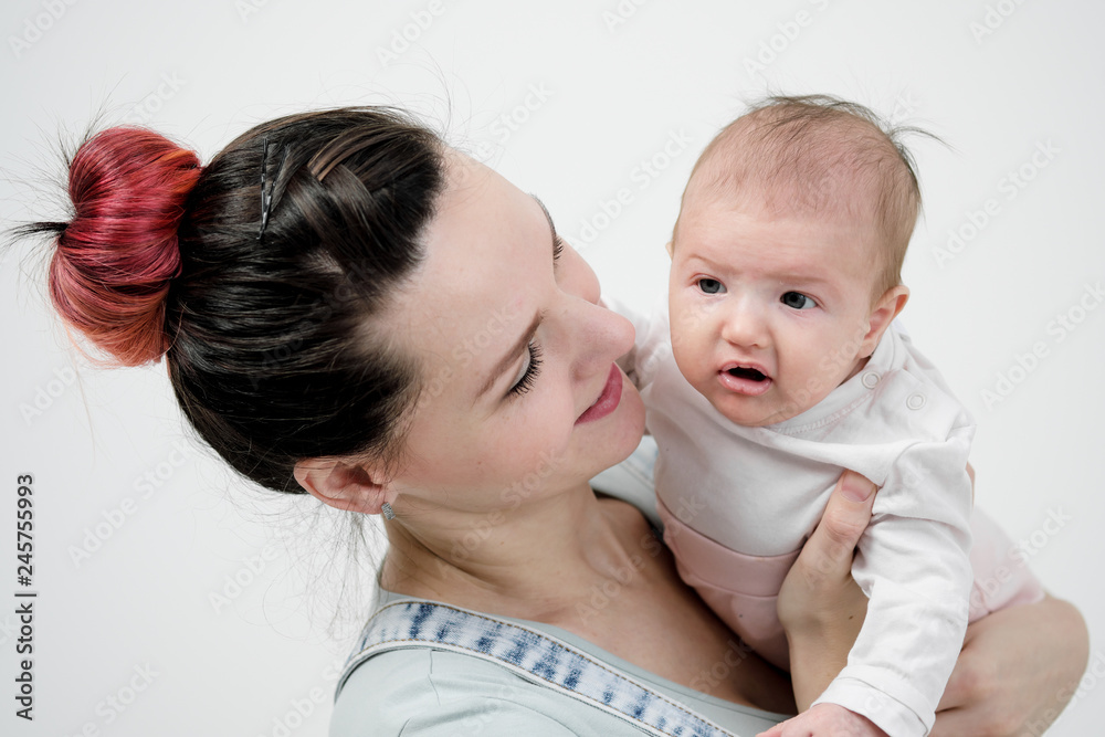 Young woman mother in denim overalls holds a baby child in her arms. White background in the studio. Young woman mother in denim overalls holds a baby child in her arms. White background in the studio.